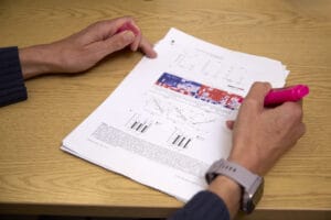 A photo of an academic paper on a desk, the text is illegible. A pair of hands are visible, this person is highlighting the paper with pink highlighter