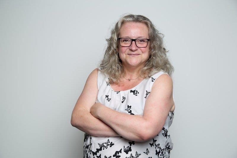 A photograph of Karen McLaren. She is in front of a plain grey background, has her arms folded and is looking at the camera smiling.